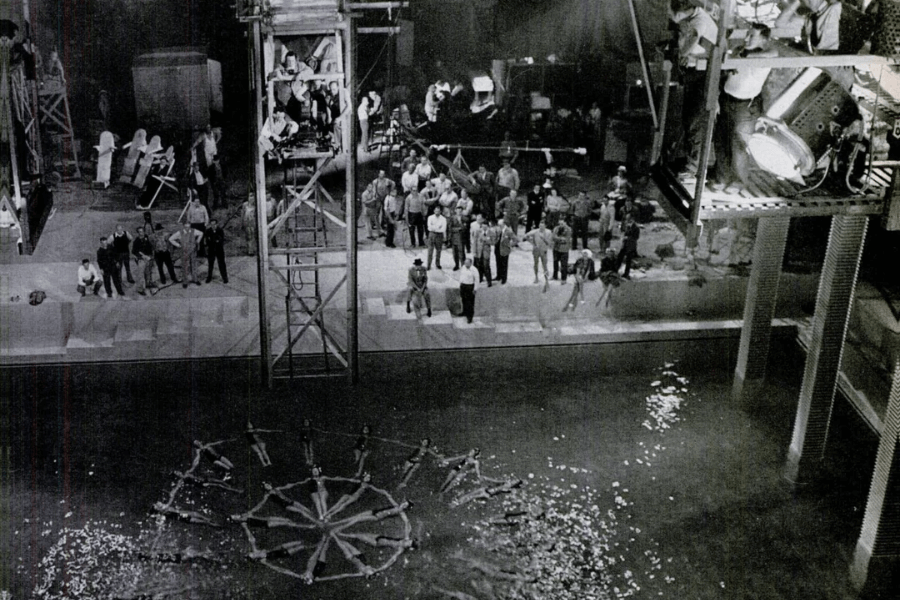 Black and white behind-the-scenes photo of a film set featuring a large synchronized swimming pool performance. A circle of swimmers forms a starburst pattern in the water, while numerous crew members, cameras, scaffolding, and massive stage lights surround the set, capturing the aquatic spectacle from above.
