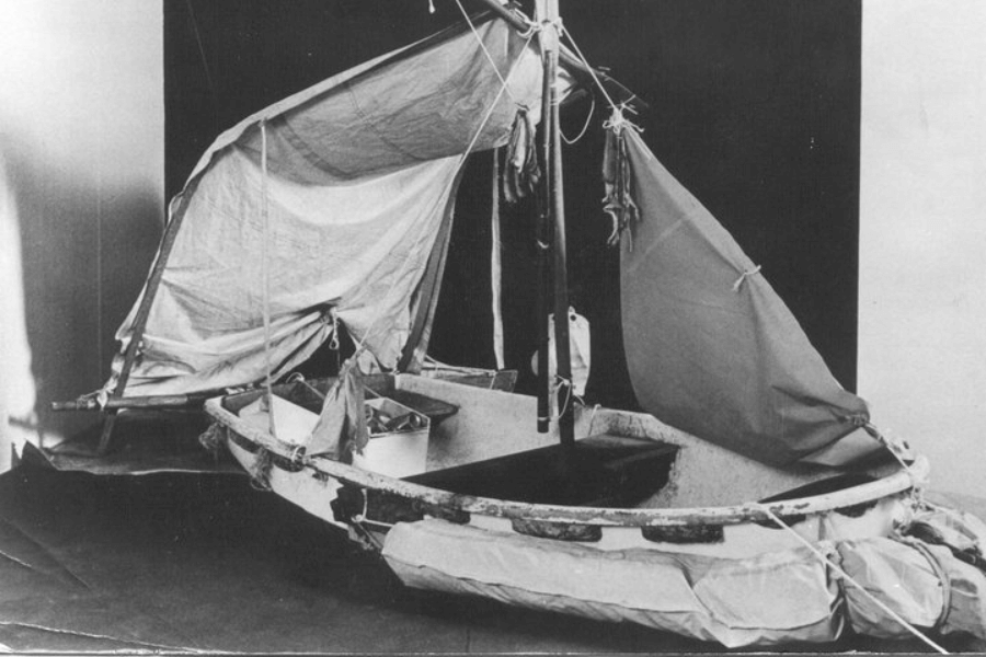 Black-and-white photo of a small, weathered lifeboat with makeshift sails and flotation collars attached along the sides. The boat is displayed indoors, outfitted with rigging, supply containers, and what appears to be hanging dried food such as turtle meat or fish.