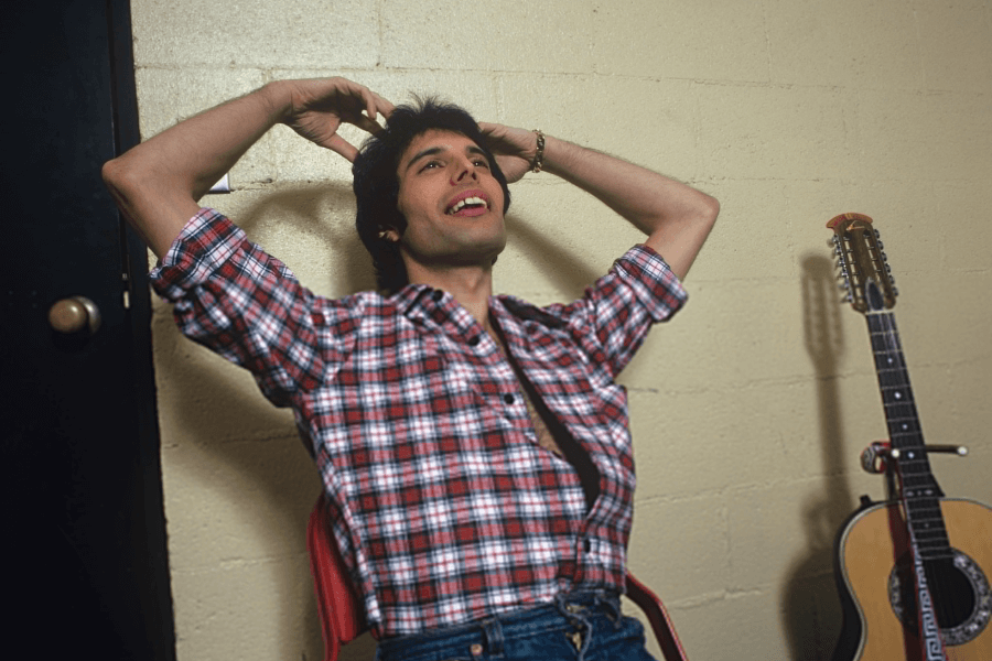 Freddie Mercury relaxing in a candid backstage moment, leaning back in a red chair with his hands behind his head, wearing a red plaid shirt and denim jeans. A 12-string acoustic guitar rests against the wall beside him, and he appears to be mid-laugh or speaking.