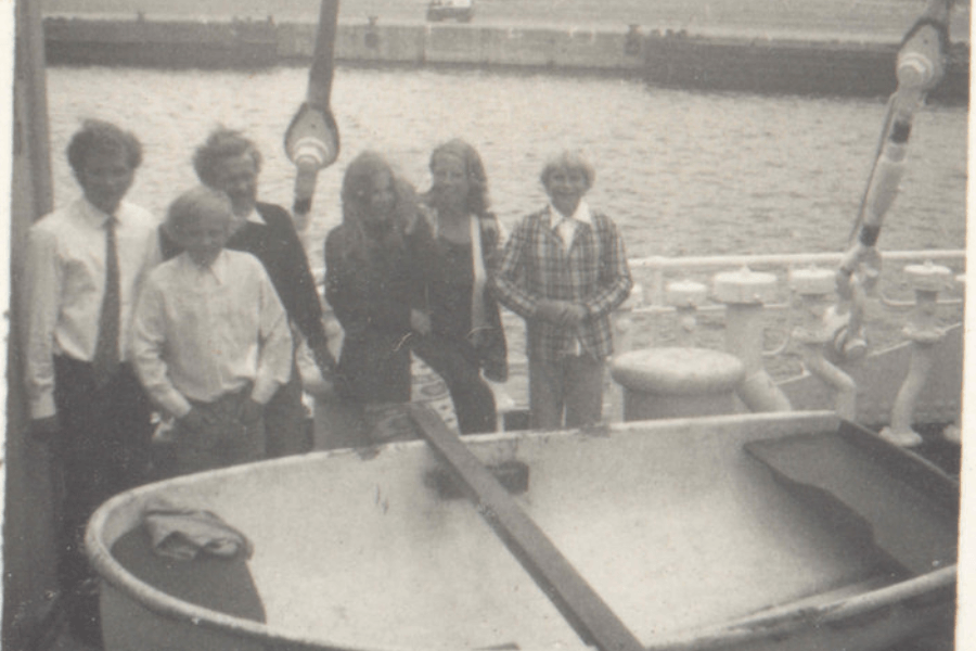 Grainy black-and-white photo of six people, including men and women, standing on the deck of a ship beside a large lifeboat. The group appears to be posing for a photo near the railing, with the harbor and pier visible in the background.