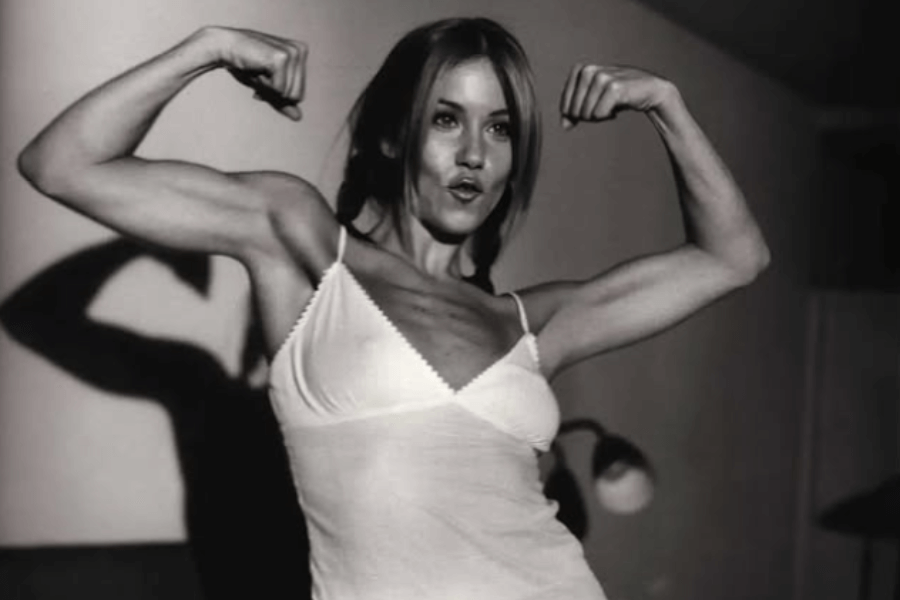 A black-and-white photo of a woman in a white camisole flexing her biceps and making a playful, determined expression. The lighting casts strong shadows on the wall behind her, emphasizing her toned muscles.