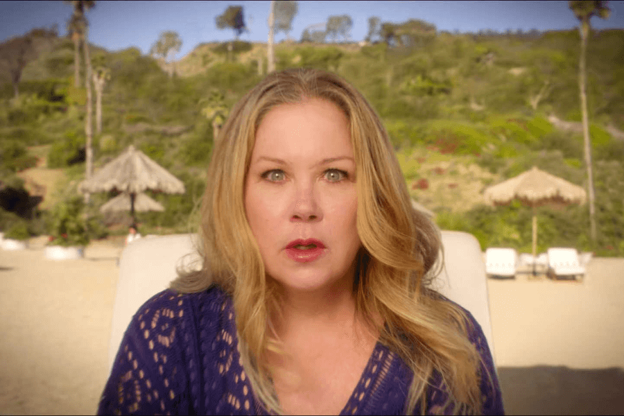 A woman with long blonde hair sits on a beach chair, looking surprised or shocked. She wears a dark blue crochet-style cover-up, with a tropical landscape, palm trees, and straw umbrellas visible in the background.