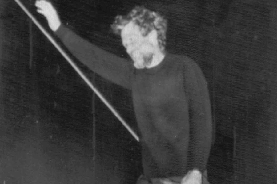 Grainy black-and-white photo of a bearded man in a dark sweater smiling as he holds onto a diagonal pole, possibly on a boat. The dim lighting and his posture suggest a candid moment, likely taken at night or in low light.