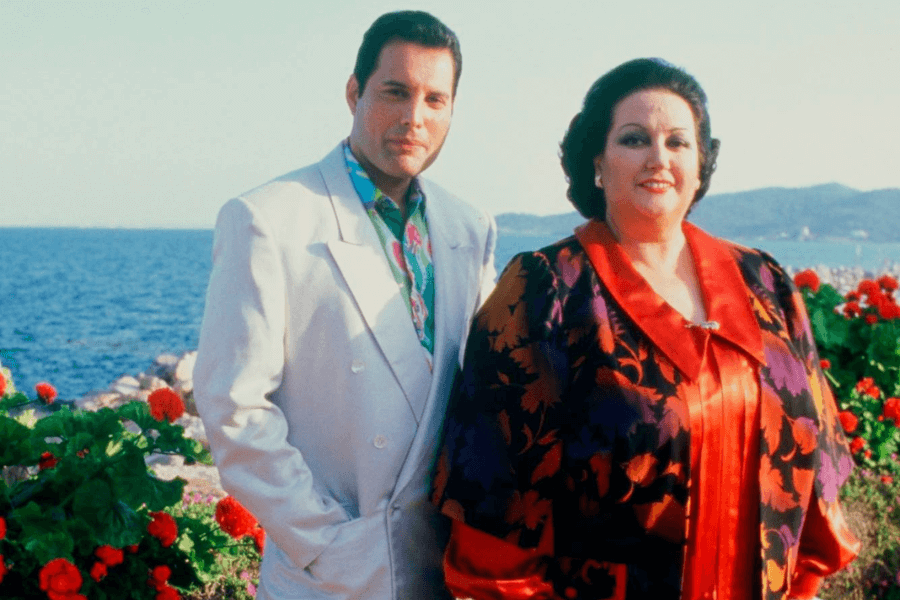 Freddie Mercury standing beside opera singer Montserrat Caballé, both dressed elegantly and posing in front of a vibrant seaside backdrop filled with red flowers. Freddie wears a white blazer over a colorful shirt, while Montserrat is draped in a bold floral-patterned outfit with a red satin collar, highlighting their famous collaboration.