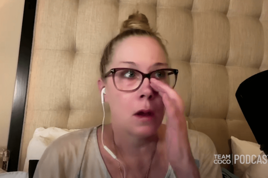 A woman with glasses and earbuds sits on a bed against a tufted headboard, appearing emotional as she wipes under her eye. She’s casually dressed and speaking into a microphone, suggesting she’s recording a podcast or interview.