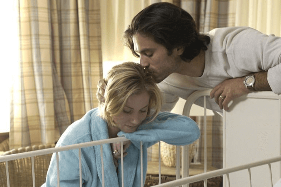 A tender moment between a couple beside a white crib: the woman, wearing a light blue robe, leans on the crib rail with a weary expression while the man gently kisses her head, offering comfort. Soft lighting and plaid curtains create a warm, intimate atmosphere.
