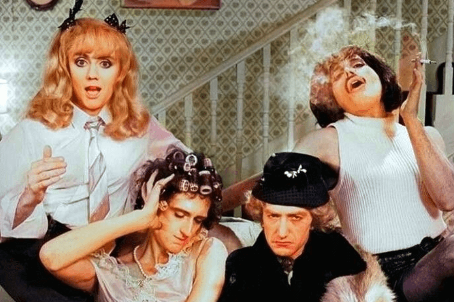 Comedic scene from Queen’s “I Want to Break Free” music video featuring all four band members dressed in exaggerated female costumes. Roger Taylor sports a schoolgirl outfit, Brian May wears curlers and a nightgown, John Deacon appears in a black hat and coat, and Freddie Mercury dramatically smokes in a sleeveless top and mini skirt, all set in a kitschy, vintage-style living room.