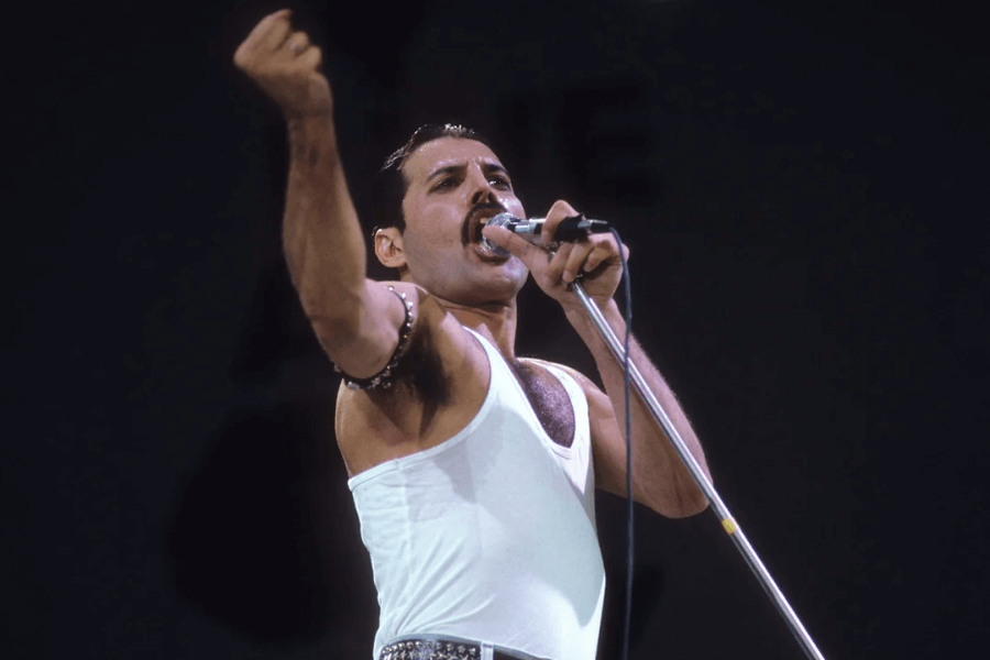 Freddie Mercury delivering a powerful performance at Live Aid in 1985, wearing a white tank top and studded belt, passionately singing into a microphone with one fist raised in the air. His dynamic stance and intense expression capture the energy of the legendary concert.