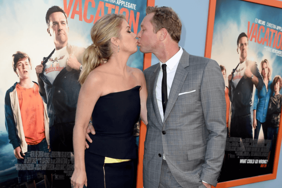 A woman in a strapless black dress and a man in a gray suit share a kiss on the red carpet in front of posters for the movie Vacation. The promotional posters behind them feature comedic poses from the film's cast, adding a playful backdrop to the affectionate moment.