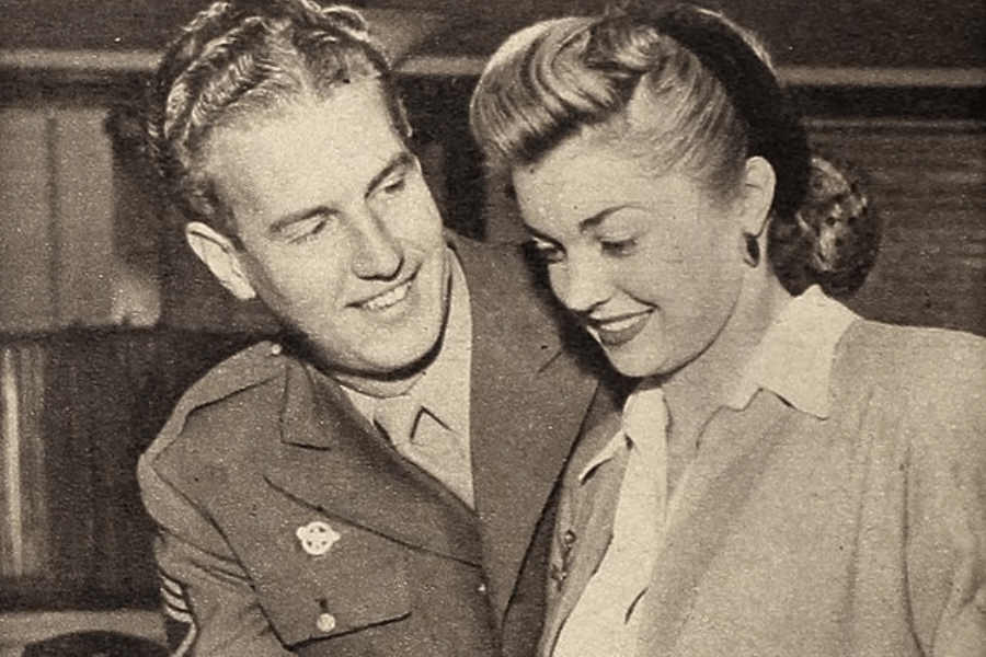 Sepia-toned image of a young couple sharing a tender moment. The man, dressed in a military uniform with visible patches and chevrons, smiles at the woman beside him. The woman wears a blouse and jacket, her hair styled in an elegant 1940s updo, and both are looking down fondly at something out of frame.