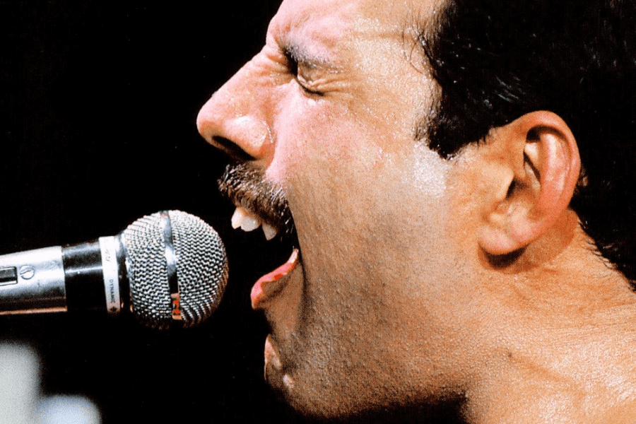 Extreme close-up of Freddie Mercury passionately singing into a microphone, eyes shut and mouth wide open mid-note, with sweat glistening on his face. The image captures the raw emotion and vocal power of his performance, highlighting his distinctive teeth and iconic mustache.