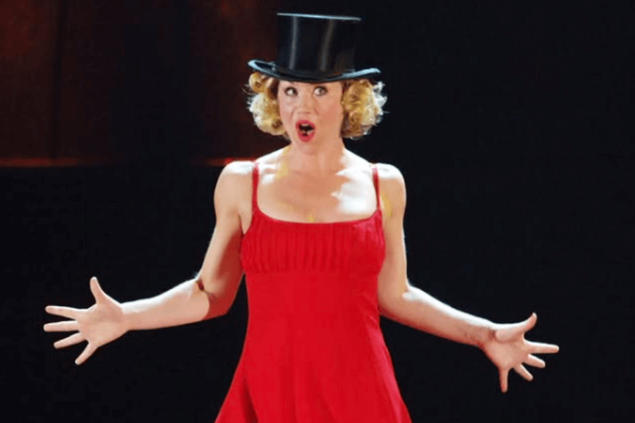 A woman in a vibrant red dress and black top hat performs dramatically on stage with an expressive face and outstretched arms. Her theatrical stance and lighting suggest a musical or cabaret-style performance.