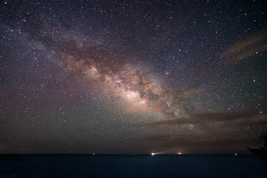 Stunning night sky photograph showcasing the Milky Way galaxy stretching across the horizon. The sky is filled with countless stars and nebulae in shades of purple, pink, and gold, with faint lights from boats or coastal sources visible along the dark ocean below.
