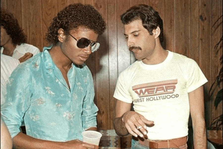 Candid photo of Michael Jackson and Freddie Mercury in conversation at a casual gathering. Michael wears a teal floral button-down shirt and aviator sunglasses, holding a drink, while Freddie stands beside him in a tight white “WEAR West Hollywood” t-shirt, holding a cigarette, both framed against wood-paneled walls.
