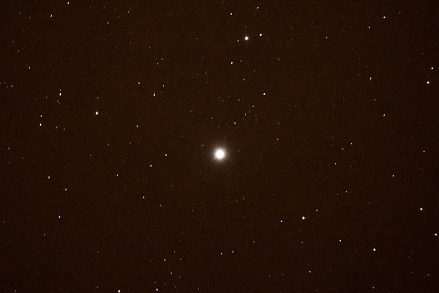 Night sky image focused on a bright celestial object—likely the planet Jupiter—surrounded by numerous smaller stars scattered across a dark brownish-black background. The central object shines prominently, outshining all nearby stars.