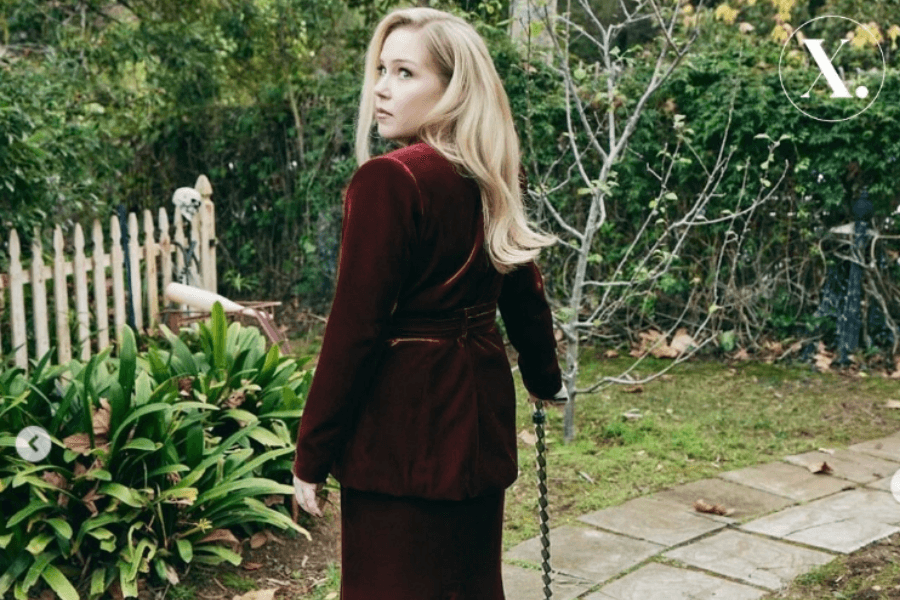 A woman with long blonde hair stands on a garden path, wearing a deep burgundy velvet outfit and looking back over her shoulder. She holds a walking cane, and the setting includes lush greenery, a white picket fence, and bare tree branches.