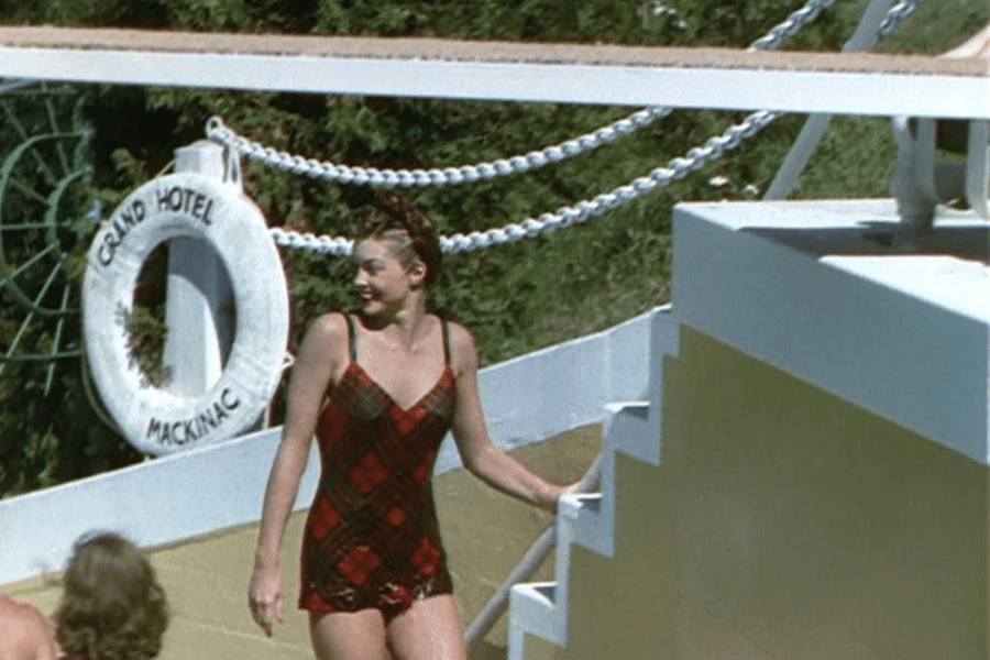 Color image of a woman in a red and black plaid one-piece swimsuit walking near a pool area. Behind her, a life preserver marked "Grand Hotel Mackinac" is mounted on a white railing, suggesting a resort or lakeside setting. She smiles while holding onto a small staircase.