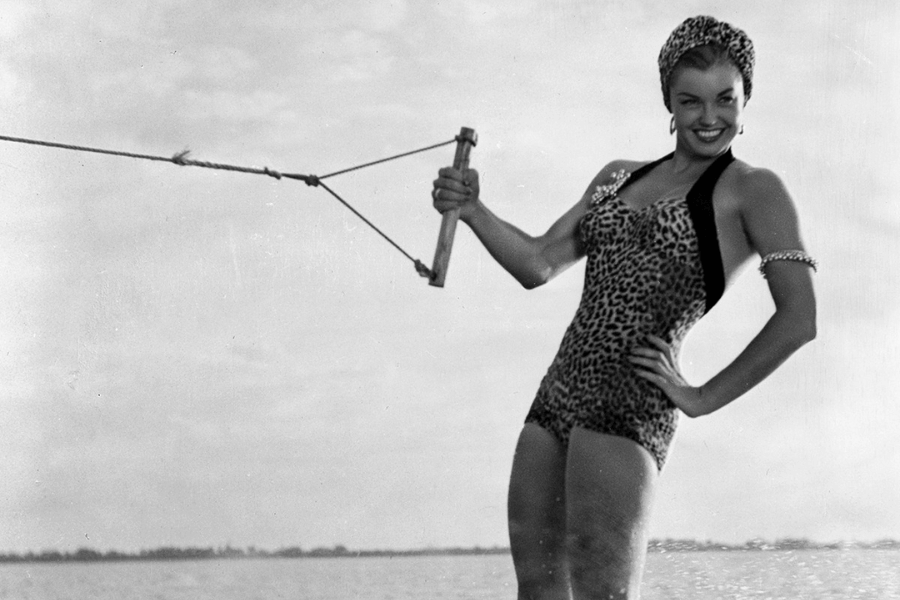 Black and white photo of a woman confidently water skiing while wearing a leopard-print swimsuit and matching headscarf. She holds a tow rope handle in one hand, smiling brightly, with one hand on her hip and water trailing behind her.