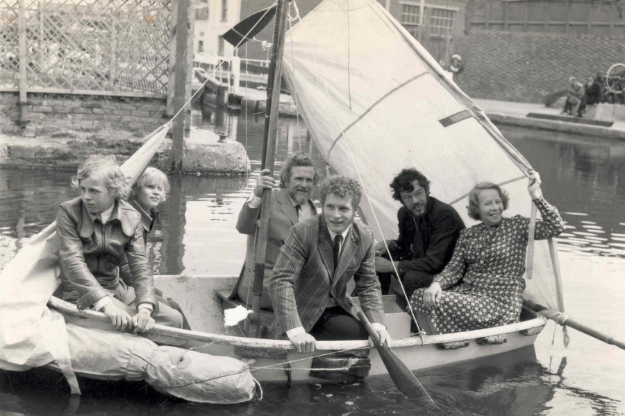 Black-and-white photo of six people—three adults and three youths—sitting in a small sailboat on a canal. The boat has a simple cloth sail and paddles, and the group appears to be dressed formally, with two men in suits and one woman in a patterned dress. Urban canal infrastructure and curious onlookers are visible in the background.