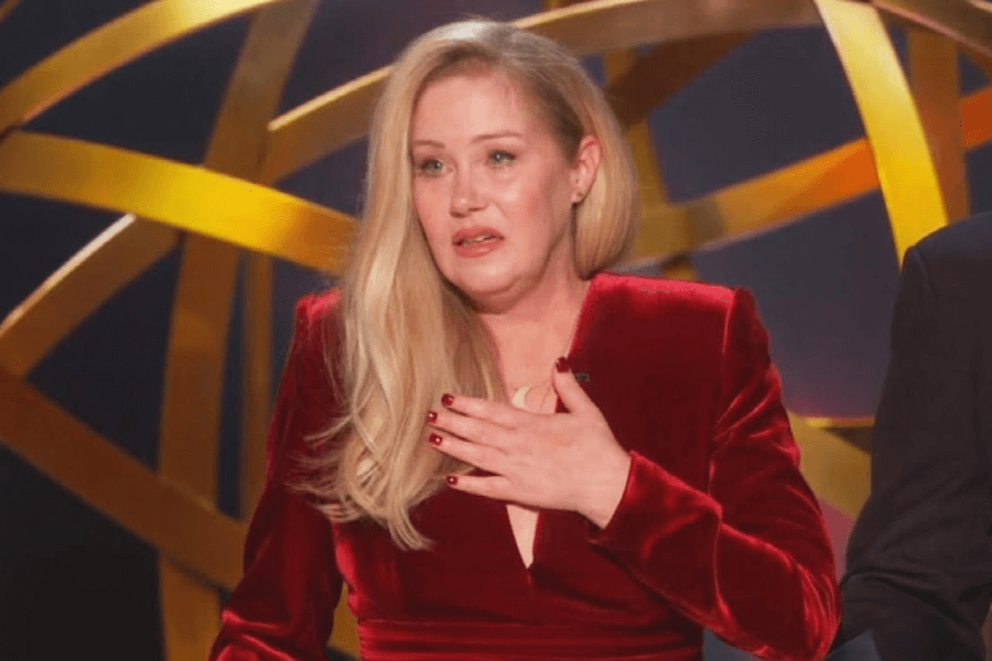 A woman with long blonde hair, wearing a deep red velvet dress, appears emotional as she holds a hand to her chest while standing on stage. The background features large, golden intertwined rings, suggesting a prestigious award ceremony setting.