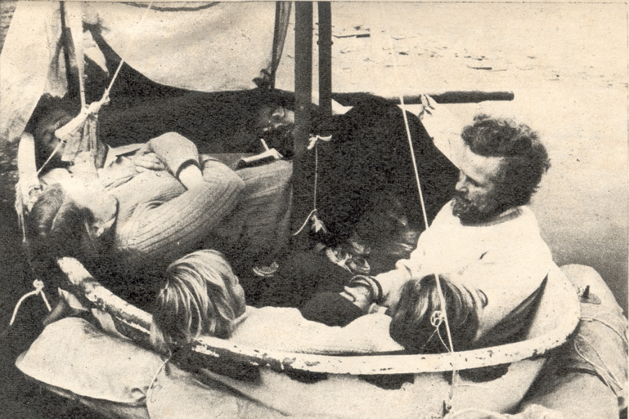 Vintage black-and-white photo of six people tightly packed into a small, weathered lifeboat. Some are lying down while others sit upright, including a bearded man in a white sweater appearing to comfort a nearby child. The scene conveys fatigue and close quarters, with ropes and a makeshift canopy rigged overhead.
