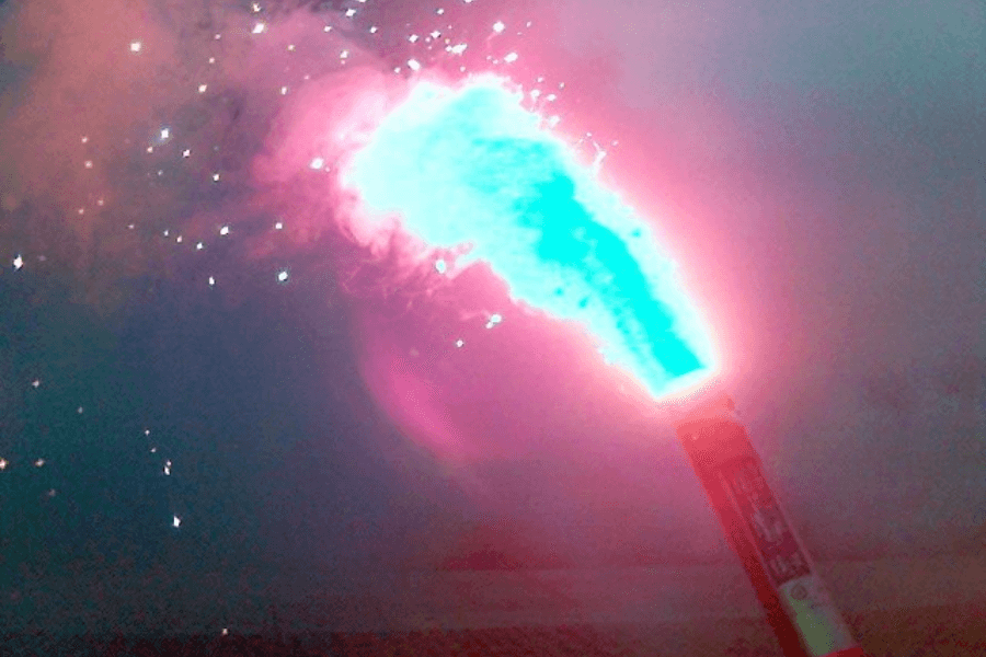 Close-up image of a handheld marine flare emitting an intense blue flame surrounded by pinkish smoke and bright sparks. The flare's cylindrical casing is partially visible, and the vivid light illuminates the surrounding area.