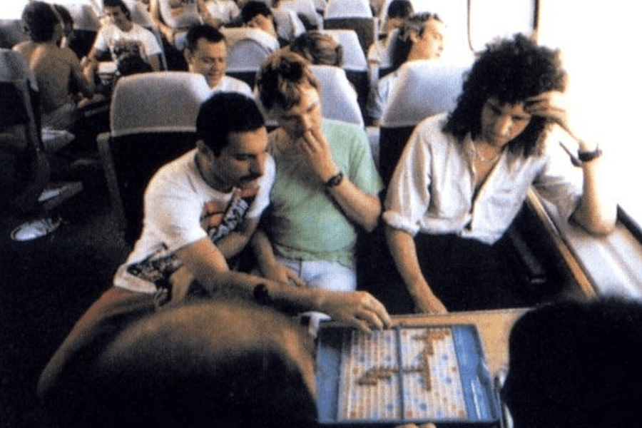 Candid photo of Queen band members Freddie Mercury, Roger Taylor, and Brian May playing a game of Scrabble on a train. Freddie and Roger are focused on the board, while Brian sits across from them looking contemplative, with other passengers visible in the background during the daylight ride.