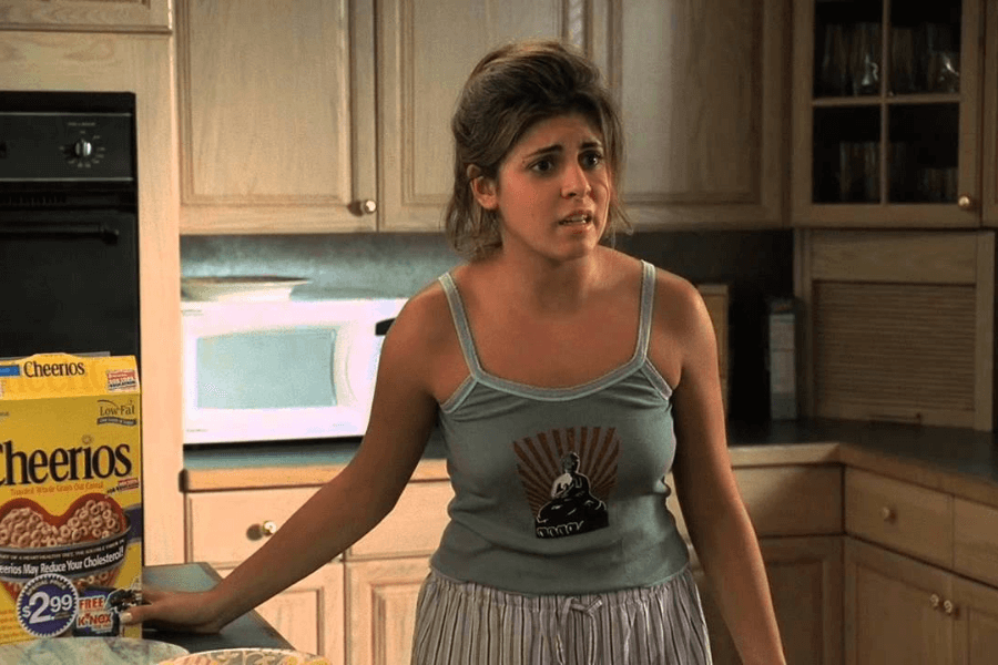 A young woman in a tank top and pajama bottoms stands in a kitchen, looking concerned or frustrated. A box of Cheerios is visible on the counter beside her, and the background features light wood cabinets and a microwave.