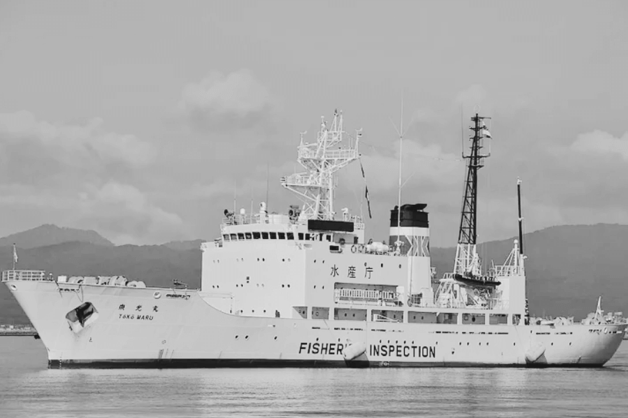 Black-and-white image of a large vessel named Tōkō Maru with “FISHERY INSPECTION” labeled in bold on the side. The ship features a tall superstructure with antennas, lifeboats, and cranes, and is docked near a mountainous coastline.