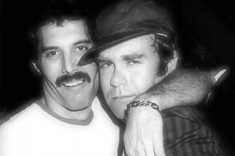 Black and white photo of Freddie Mercury smiling with his arm around Elton John, who wears a cap and a striped jacket. The two music legends are captured in a candid, close moment, highlighting their friendship and camaraderie.