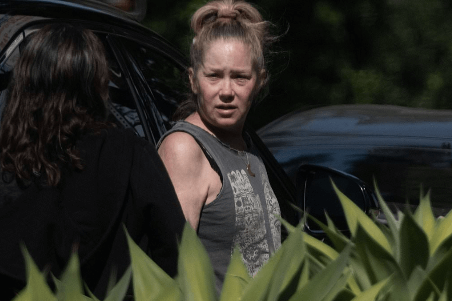 A woman with a messy bun and sleeveless graphic tank top stands outdoors beside a dark car, speaking with another person whose back is to the camera. Green foliage partially obscures the lower part of the image.