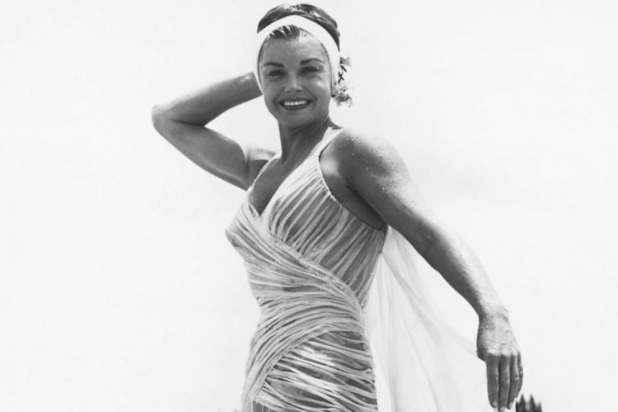Black and white photo of a woman in a sheer, ruched swimsuit with a flowing wrap, posing confidently with one arm behind her head. She’s smiling brightly and wearing a headband, likely poolside or at the beach, with water droplets visible on her skin.