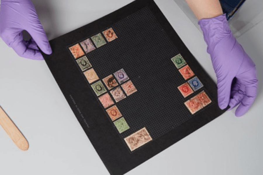A pair of gloved hands carefully handle a black stamp album page displaying a colorful assortment of vintage postage stamps arranged in a staggered grid. The stamps vary in size and design, set against a fine mesh background, suggesting archival preservation or collection curation.