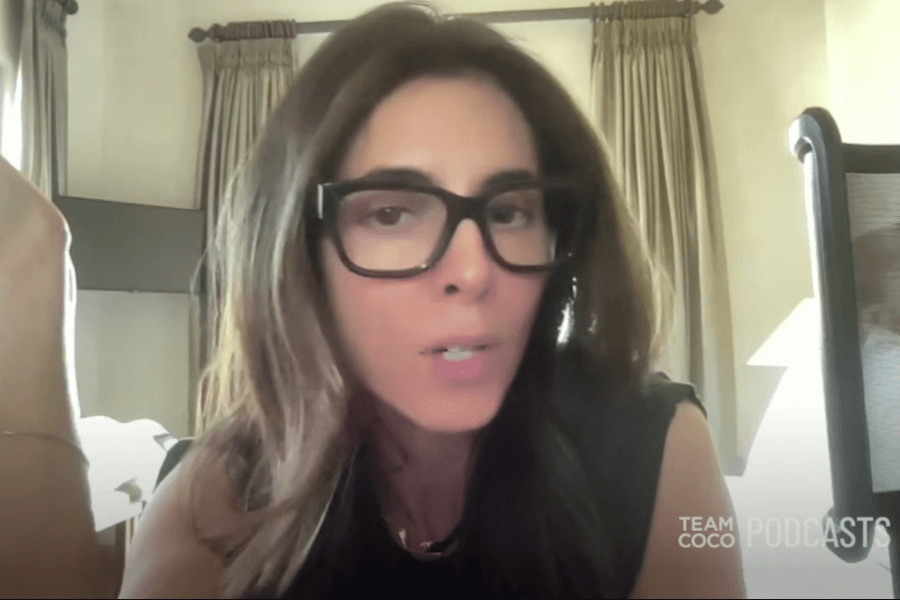 A woman with long brown hair and oversized black glasses speaks during a podcast recording, sitting in a softly lit room with beige curtains and a microphone nearby. The “Team Coco PODCASTS” logo is visible in the lower right corner.