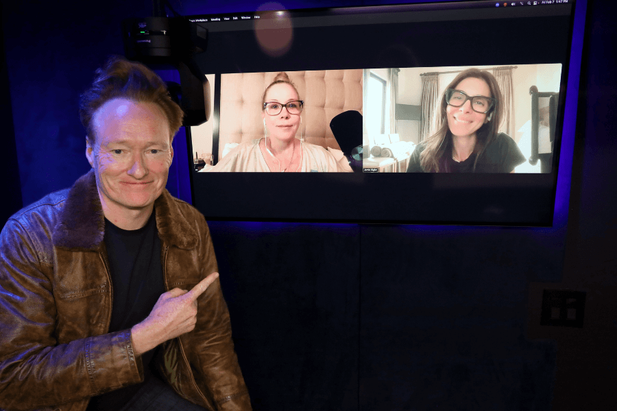 Conan O'Brien sits in a dark recording studio wearing a brown leather jacket, smiling and pointing toward a screen displaying two women on a video call. The women, both wearing glasses, appear to be participating in a podcast session, with one seated against a tufted headboard and the other in a curtain-lined room.