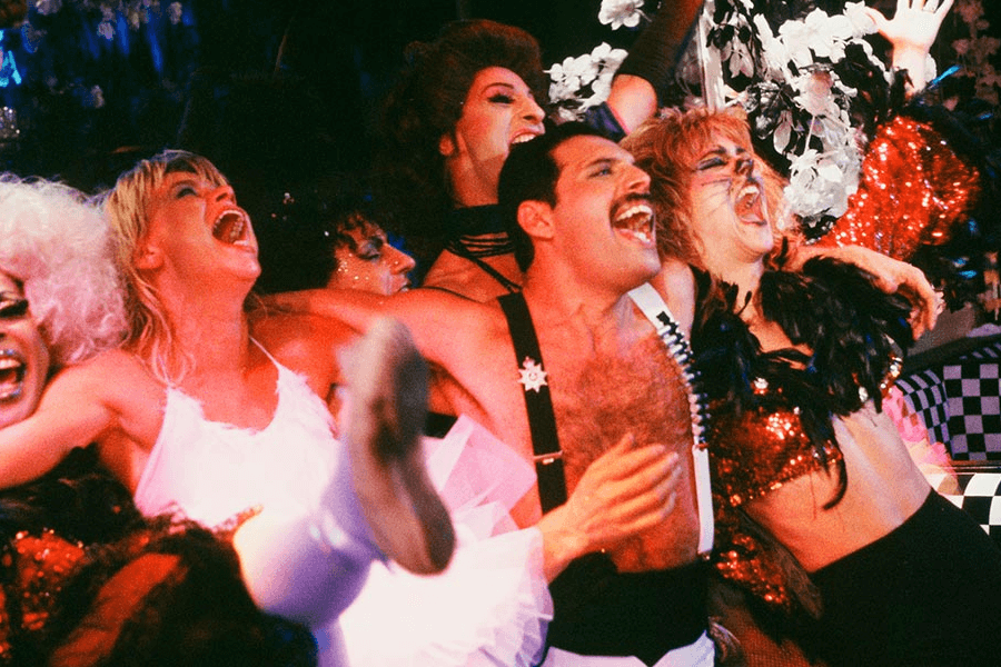 Freddie Mercury celebrating exuberantly at a lively party, surrounded by drag queens and costumed performers in extravagant makeup, glitter, and vibrant outfits. With arms around each other, they sing and laugh amid dramatic lighting and flamboyant décor, capturing the spirit of 1980s nightlife.