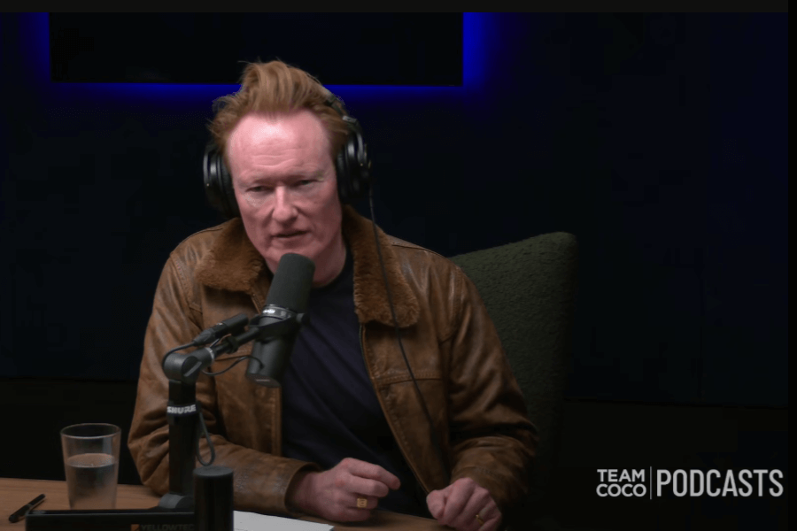 Conan O'Brien records a podcast in a dimly lit studio, wearing headphones and a brown leather jacket. He speaks into a professional microphone with a glass of water, papers, and the “Team Coco PODCASTS” logo visible on the table.