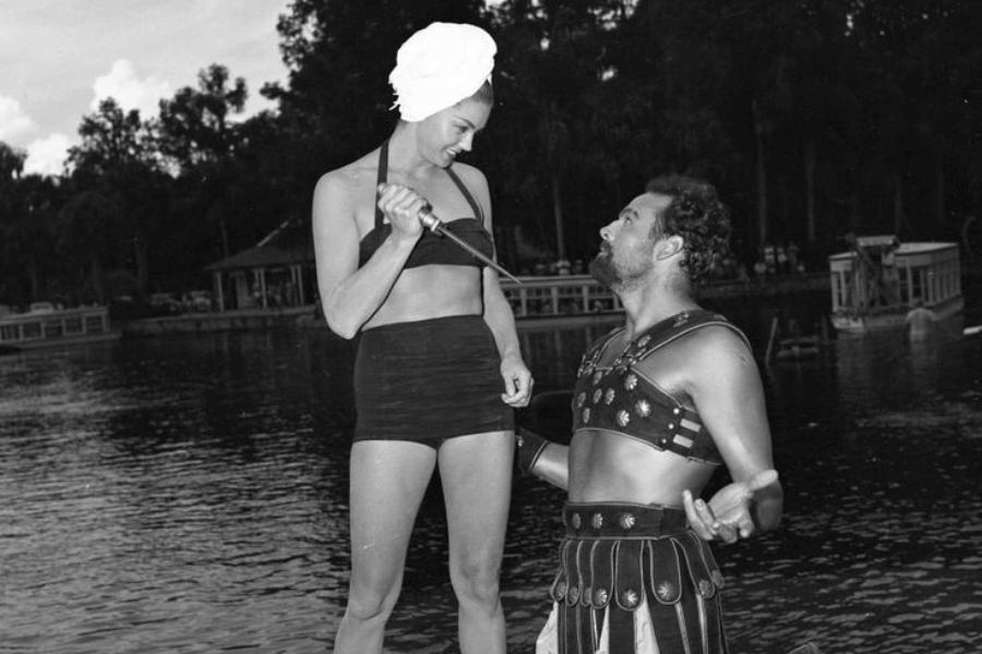 Black and white photo of a playful scene by the water, featuring a woman in a high-waisted two-piece swimsuit and a towel wrapped around her head, holding a prop sword toward a man dressed in Roman gladiator attire. The man kneels before her with an exaggerated gesture, while both appear to be enjoying the humorous moment.