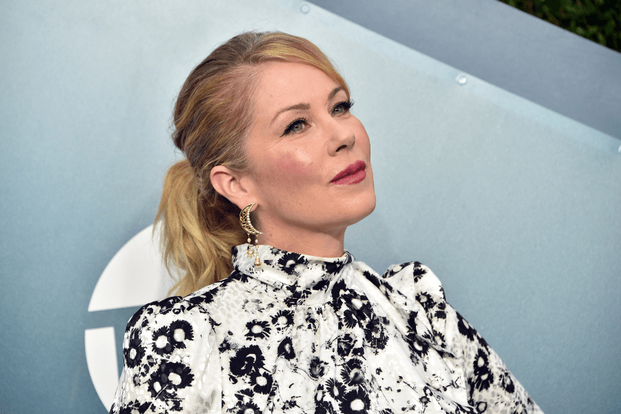 A woman with blonde hair tied in a low ponytail poses in front of a blue-gray backdrop, wearing a high-neck white and black floral dress with statement earrings. She gazes upward with a composed expression, highlighting her elegant makeup and styling.