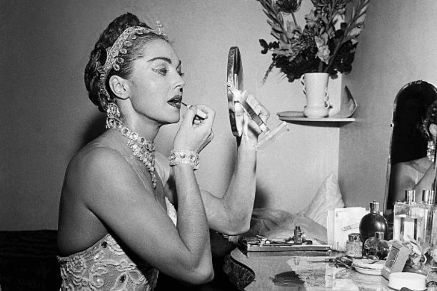Black and white photo of a glamorous woman applying lipstick while looking into a handheld mirror at a vanity. She wears a sparkling gown with ornate jewelry including a bejeweled headband, necklace, and bracelet. The vanity is cluttered with perfume bottles and makeup, with a vase of flowers in the background.