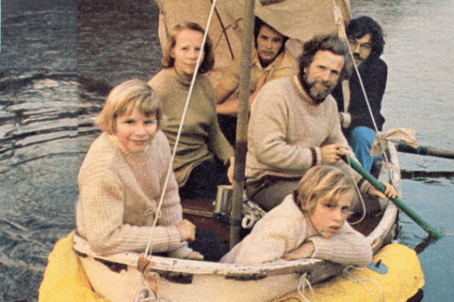 Color photo of six people sitting in a small, weathered lifeboat with a yellow flotation collar. Two children in cream-colored sweaters are at the front, smiling and leaning on the boat’s edge. Behind them sit a woman, three men, and another child, with makeshift sails and supplies visible onboard, suggesting a reenactment or post-rescue moment.