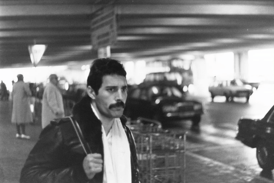 Black and white candid photo of Freddie Mercury walking through a dimly lit parking garage or terminal, wearing a leather jacket and carrying a bag over his shoulder. His expression is serious and focused, while blurred cars and people in coats move in the background.
