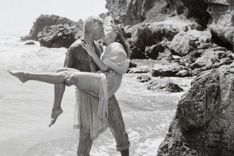 Black and white romantic photo of a man standing in shallow ocean water, holding a woman in his arms as they share a kiss. Both are wet from the sea; the man wears a button-up shirt and rolled-up trousers, while the woman is in a light, flowing dress. They are surrounded by rugged coastal rocks.