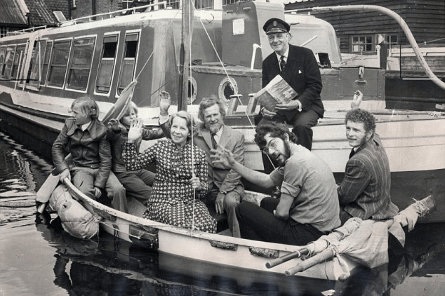 Black-and-white photo of seven people sitting in a small lifeboat, smiling and waving at the camera. A uniformed man holding a book sits at the stern, while the rest—including a woman in a patterned dress and men in casual or formal attire—are arranged along the sides. A larger boat is docked behind them.