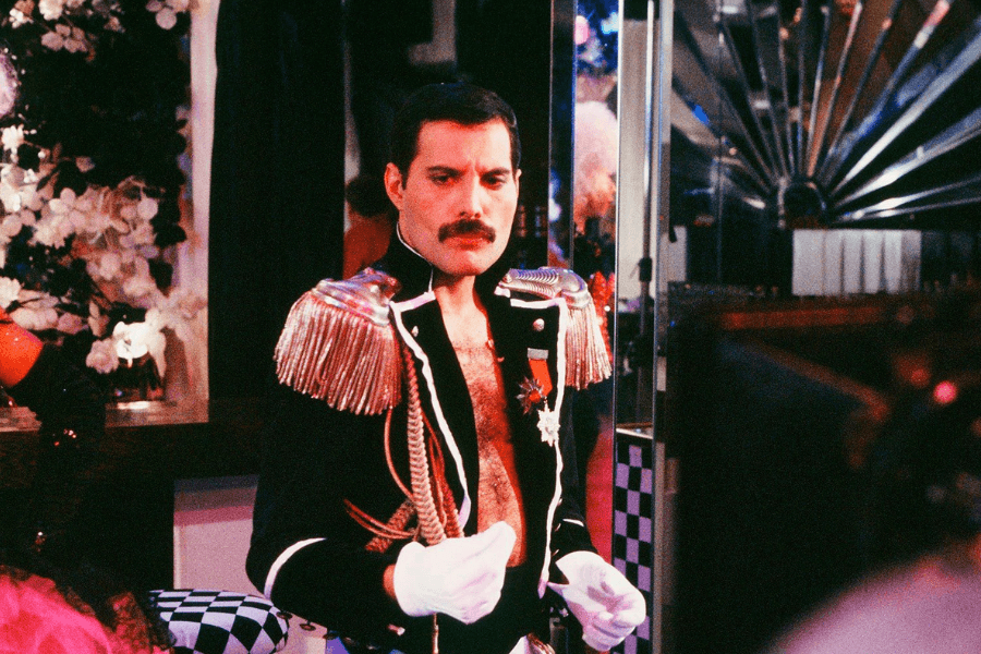 Freddie Mercury in a flamboyant military-style costume with gold fringe epaulettes, medals, and a deep V-neck jacket revealing his chest, standing confidently in a room filled with mirrors, floral decor, and black-and-white checkered patterns. The vibrant colors and theatrical setting suggest a party or performance environment.