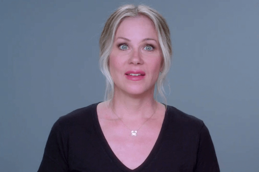 A woman with light blonde hair styled in a soft updo stands against a plain gray background, wearing a black V-neck top and a delicate necklace with a clover-shaped pendant. She looks directly at the camera with a composed expression.