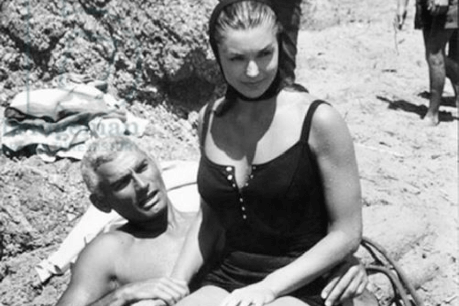 Black and white beach photo of a man reclining on a towel, holding a woman who is sitting on his lap. The woman wears a vintage black swimsuit with buttons and a headscarf, gazing off to the side with a subtle smile. The sandy shoreline and other beachgoers are visible in the background.