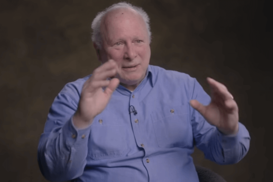 An elderly man with a bald head and white hair is seated, wearing a blue button-down shirt and gesturing with both hands while speaking. A lapel microphone is clipped to his shirt, indicating he's being interviewed, and the background is softly lit in a warm brown gradient.