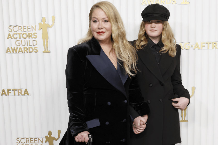 At the Screen Actors Guild Awards, a woman in a black velvet coat with navy satin lapels stands holding hands with a young girl dressed in a matching black outfit and newsboy cap. Both have long blonde hair and pose confidently on the red carpet.