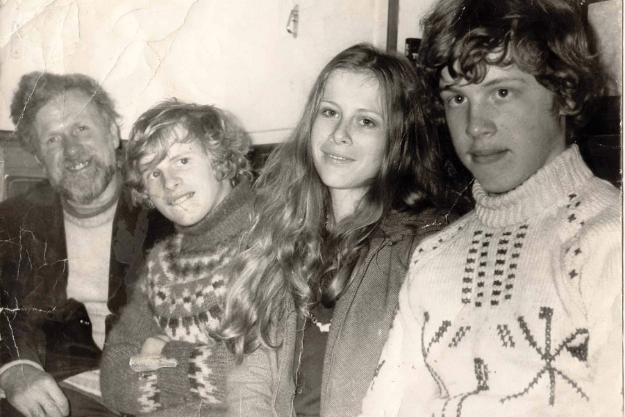 A black-and-white photo of four people sitting closely together indoors, smiling gently at the camera. From left to right: a bearded man in a sweater and blazer, a young person in a patterned wool sweater, a young woman with long hair, and another young person in a white knit sweater. The image has visible creases, giving it a nostalgic, worn appearance.